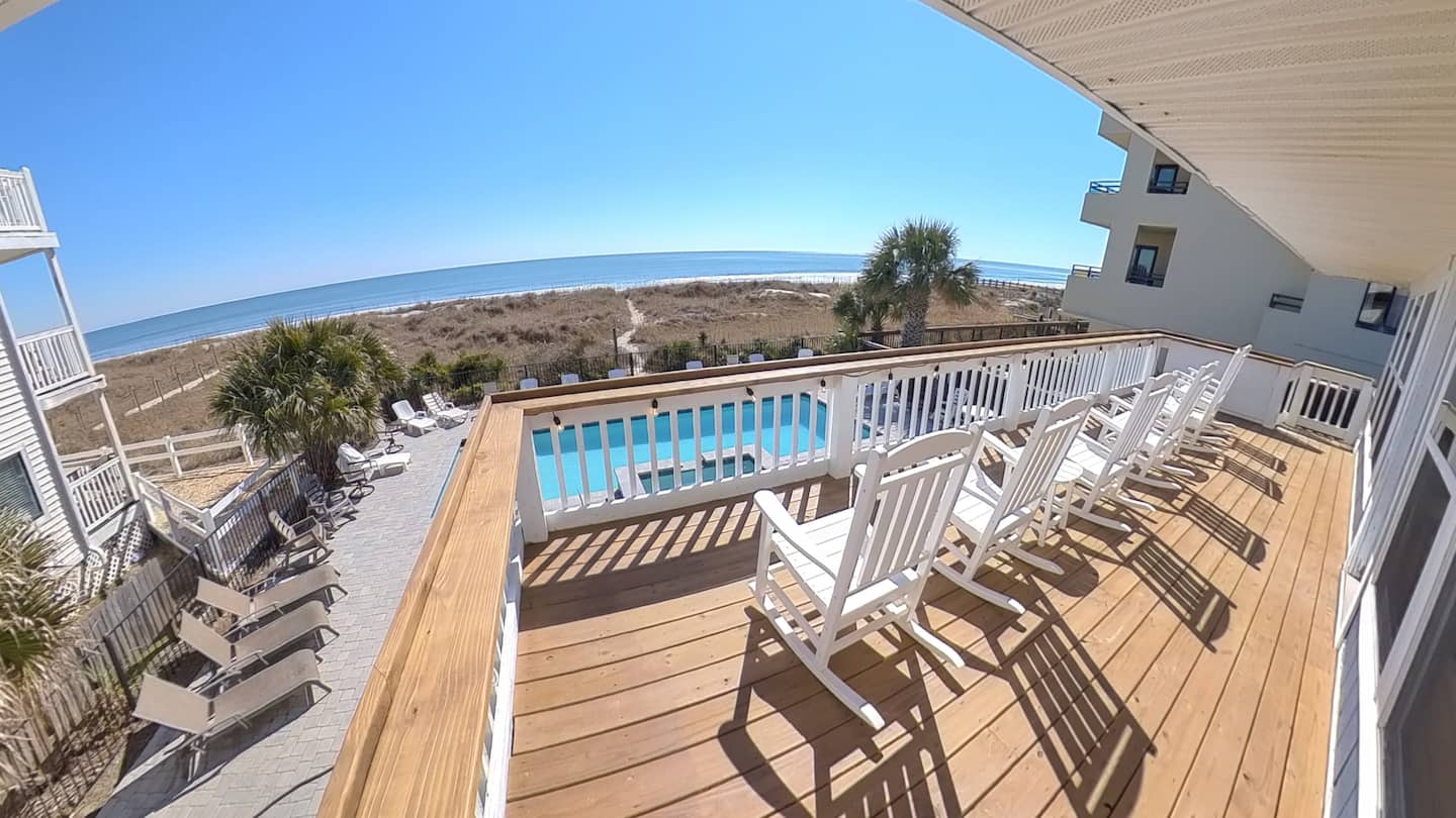 White chairs on an elevated wooden deck overlooking a swimming pool, beach, and ocean.