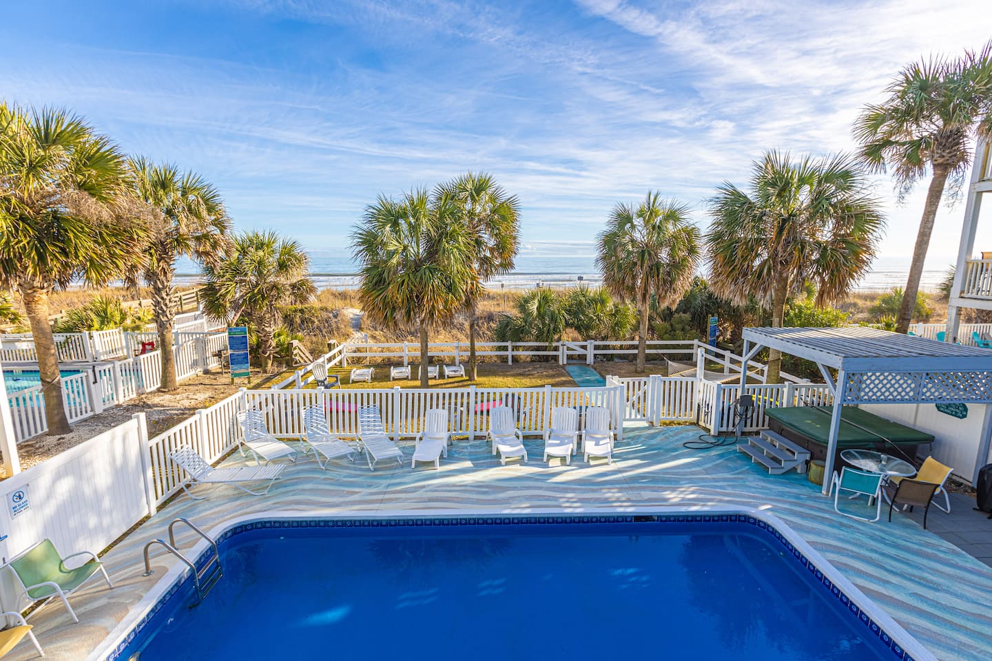 Swimming pool bordered by white fencing, loungers, and palm trees.